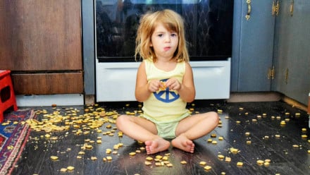 A little girl sitting on the floor surrounded by spilled cereal which she is eating