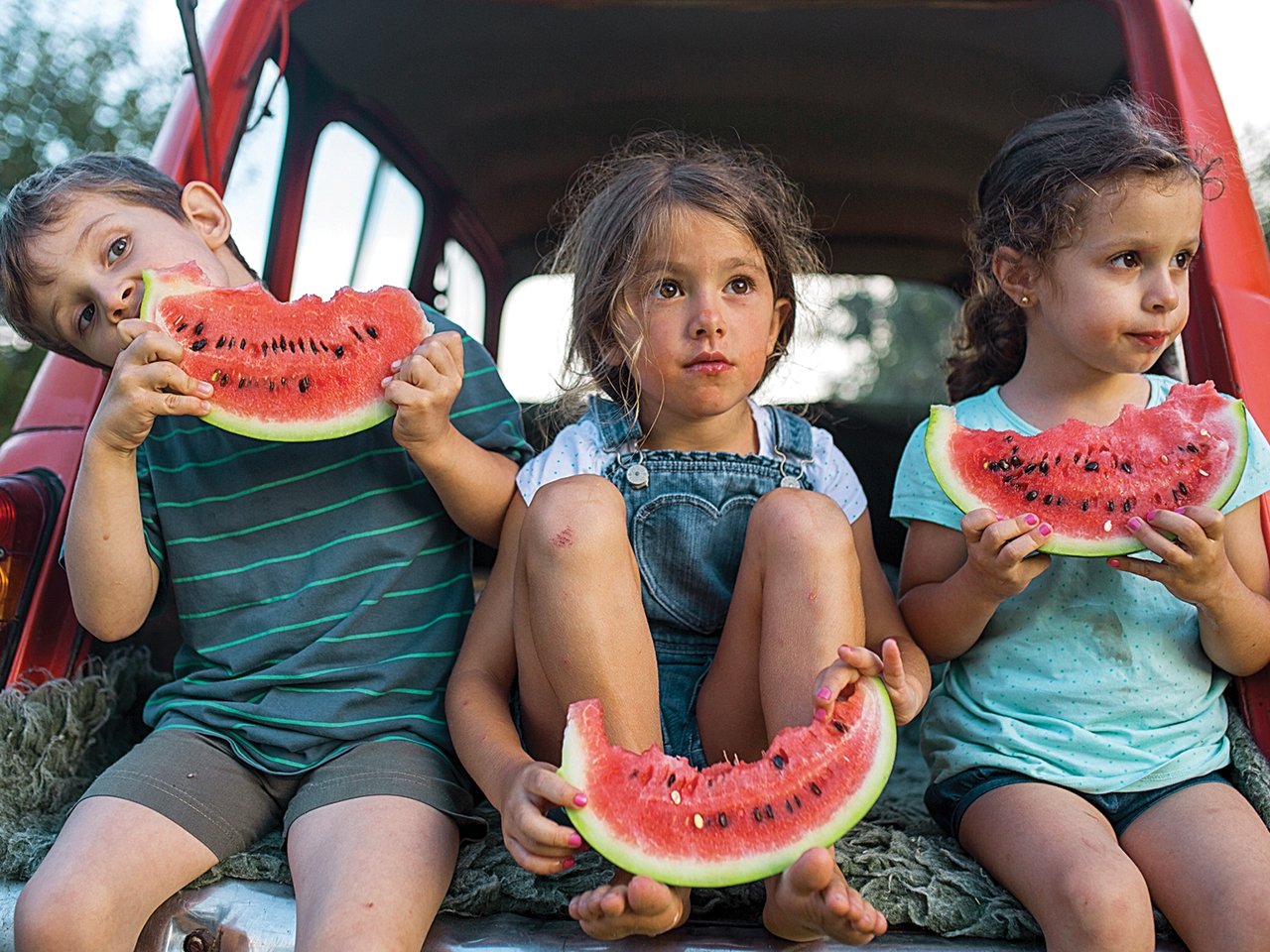 Three kids eating watermelon in the trunk of a car