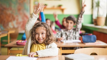 A little girl with curly blonde hair in a yellow top and white sweater raising her hand at her desk in class
