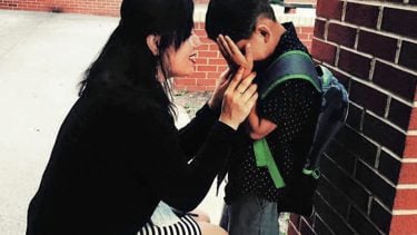 A little boy wearing a backpack and crying into his hands to his mom
