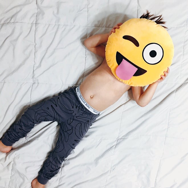 A kid lies on a bed and covers his face with a winking tongue emoji pillow