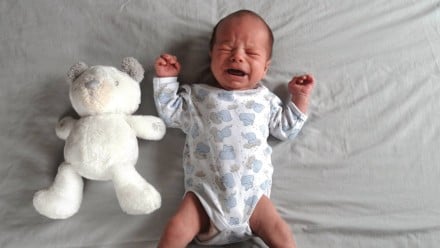 A baby crying on a sheet next to a teddy bear