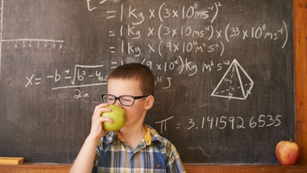 little boy with glasses eating an apple in front of a chalkboard with math on it