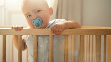 baby standing up in a crib with a soother looking grumpy