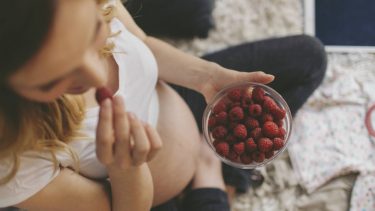 A pregnant woman sitting on the bed eating a bowl of raspberries