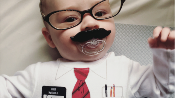 Baby in a suit onesie wearing glasses and a mustache pacifier