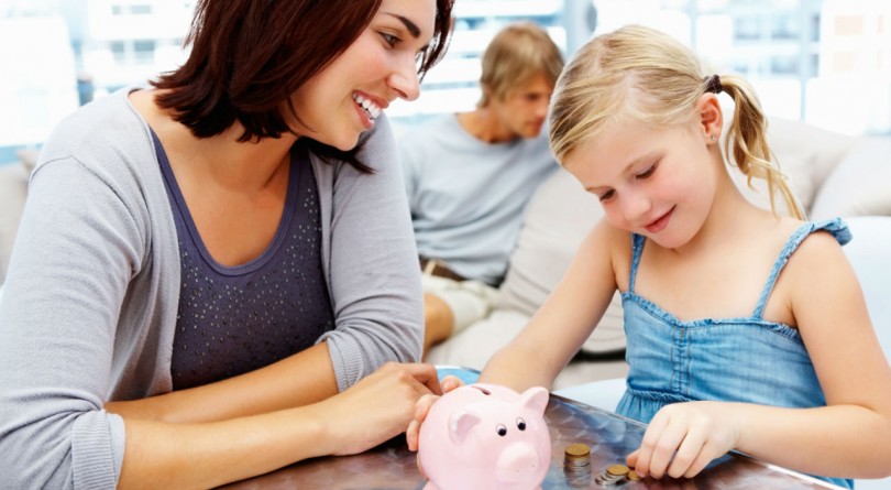 Mom and daughter sit at table while daughter puts her allowance into her piggy bank