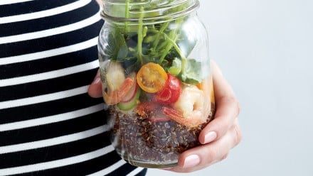 pregnant woman holding mason jar filled with red quinoa, shrimp, cherry tomatoes and spring mix