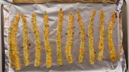 A row of asparagus with breading on a tinfoil lined baking pan