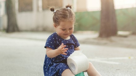 A little girl in a dress sitting on a potty and holding toilet paper