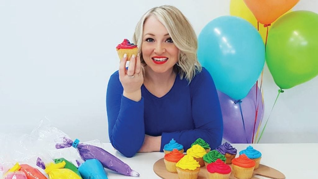 Author holding up a cupcake and sitting at a table with colourful cupcakes and balloons