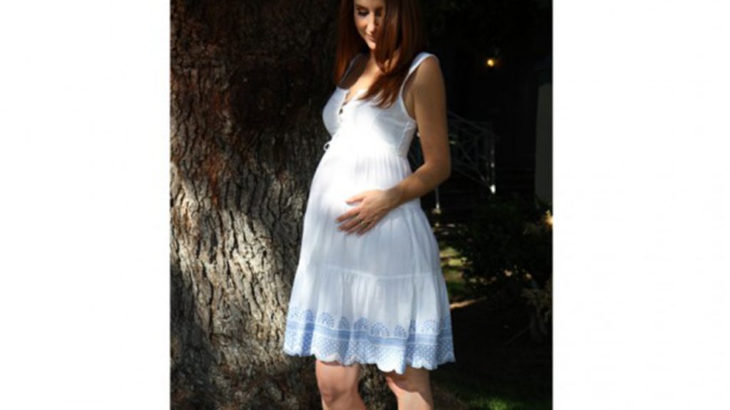 Pregnant woman stands by a tree in a white dress