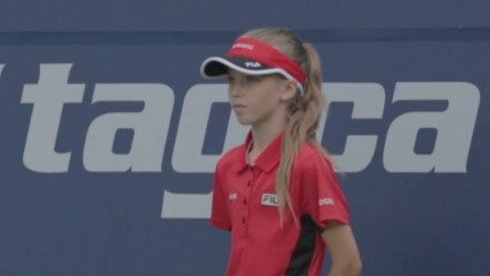 A little girl on a tennis court wearing a visor and red shirt