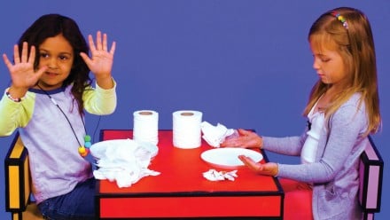 Two kids sitting at a red table with toilet paper on it