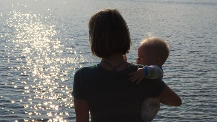 woman stands on dock holding baby and holding hand of little girl