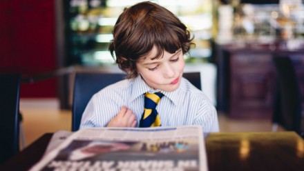little boy wearing a dress shirt and tie reading a newspaper