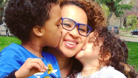 Mother wearing bright blue glasses gets kisses on the cheek from her two kids