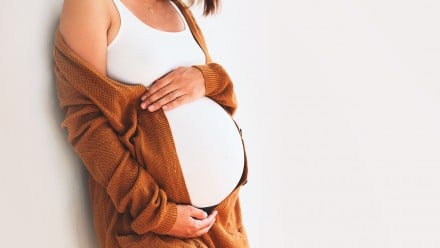 Pregnant woman in a burnt orange cardigan leans against a wall holding her belly