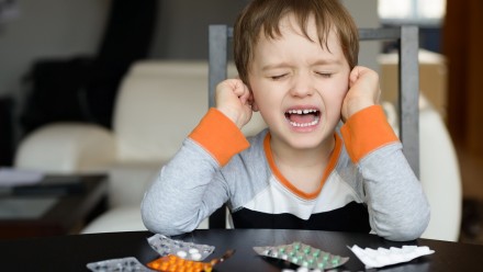 Kid sitting at a table with meds in front of him. He is holding his ears and crying