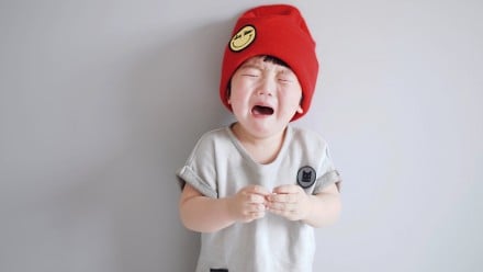 Little kid wearing bright red beanie and a grey shirts stands in front of a wall crying