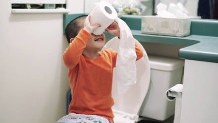 A little boy sitting on the toilet looking through toilet paper