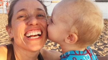 Toddler son gives his mom a kiss on the cheek as they sit on the beach
