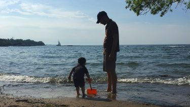 Dad looks over his son as they stand in the waves at the lakeshore. the son is digging into the sand with a red shovel