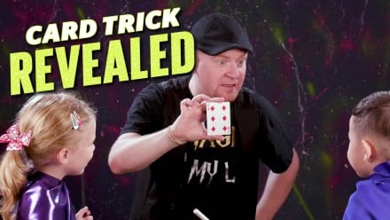 A magician showing a card to two kids wearing capes