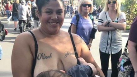 A woman breastfeeds her baby outside while two other women behind her scowl.
