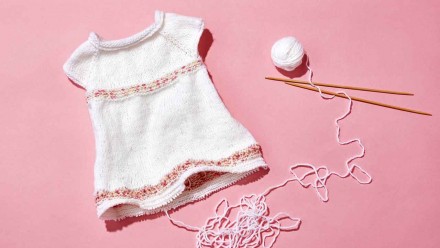 White and pink knit baby dress with knitting needles and ball of yarn