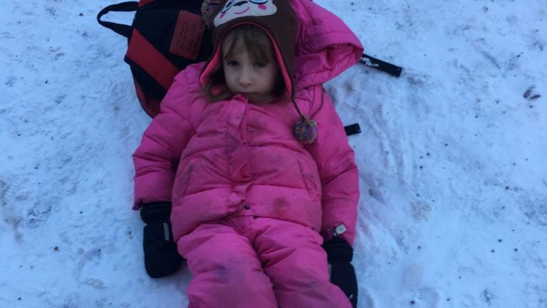 A little girl in a pink snowsuit lies on the floor with her head against her backpack.