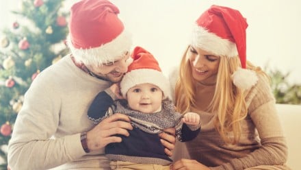 Christmas photo of a happy family of three with their newborn baby around a decorated Christmas tree