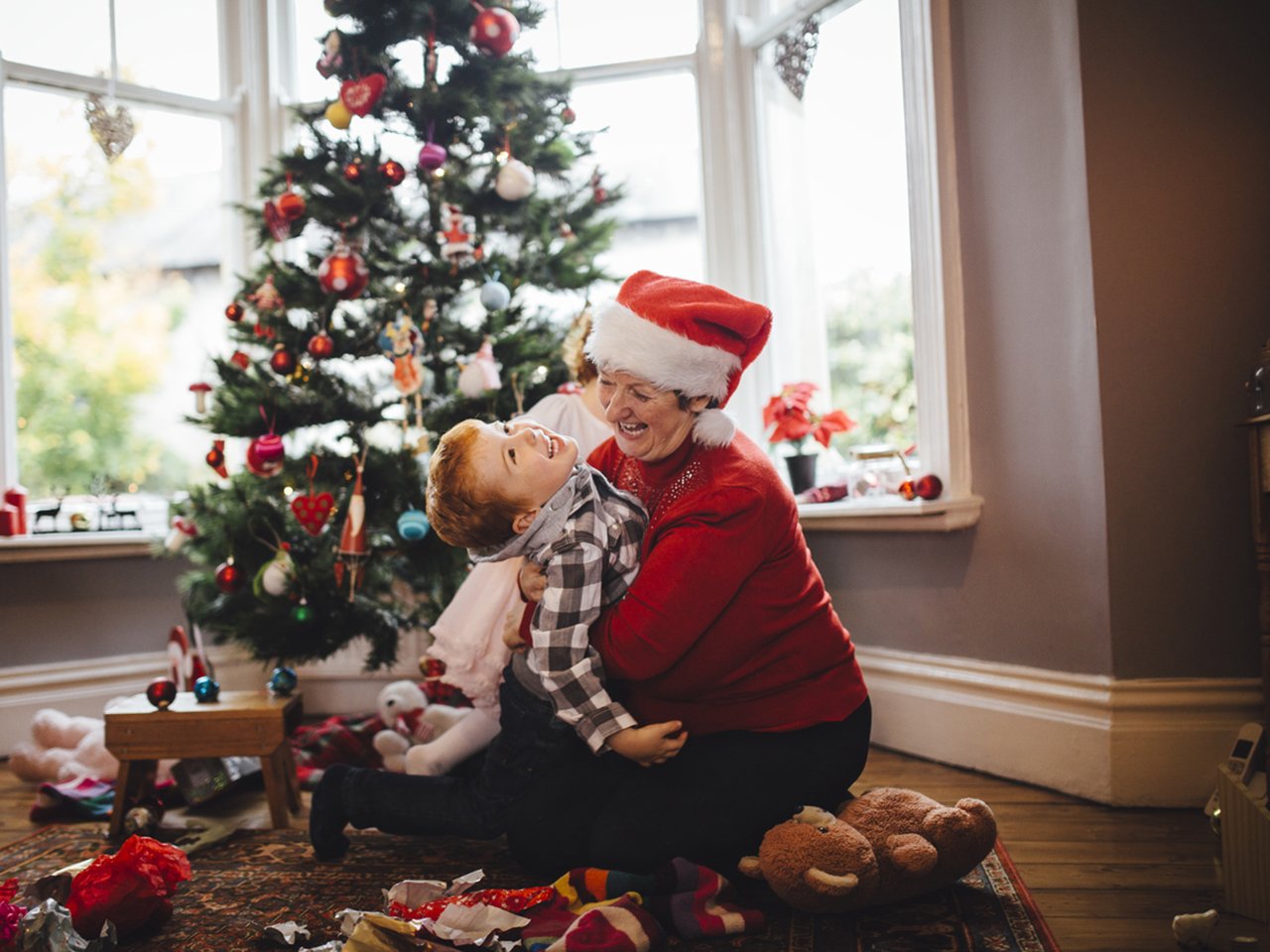 A grandmother wearing a santa hat and her grandson are celebrating Christmas morning together. The elderly woman is sitting on the floor with the little boy and holding him in a tight embrace as they play. A Christmas tree can be seen in the background and toys scattered around the floor