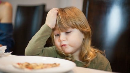 A young girl pouts at a plate of food on the table in front of her.