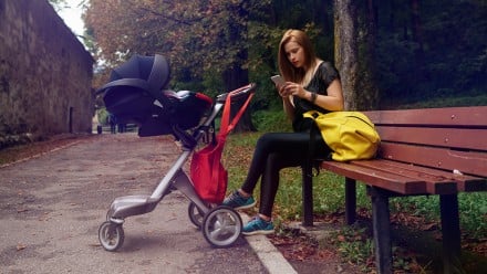 Young mother texting on her phone, sitting on a park bench with her baby in a stroller