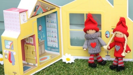 Two small wooden elves dressed in knitted clothes stand in front of a small cardboard play-house.