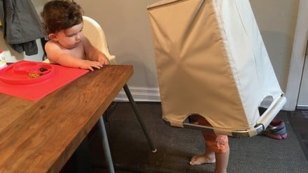 Kid playing with laundry hamper while baby eats at the table in a high chair