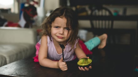 An unhappy child holds an avocado and a spoon