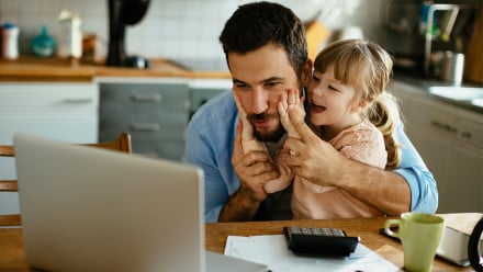 A man tries to work on a laptop while his daughter grabs his face