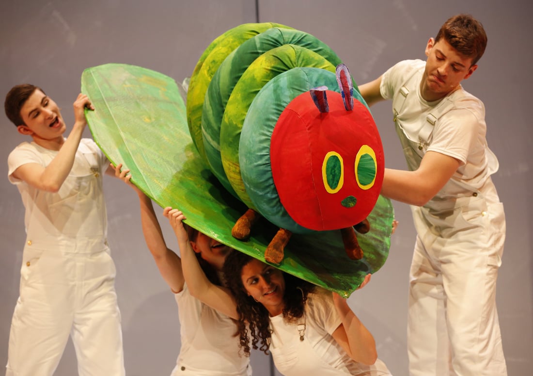 Three actors in white manipulating a caterpillar puppet on a leaf