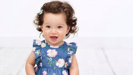 A baby in a blue flower dress smiles at the camera