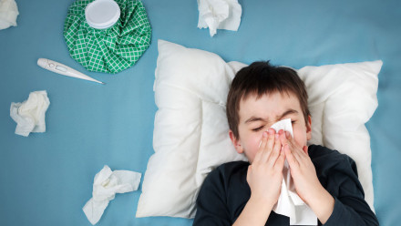 Little boy with cold and flu blowing his nose while lying down on bed surrounded by tissues, thermometer and heat pack