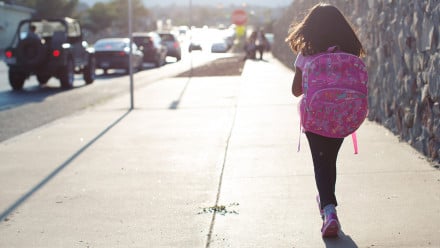 A young girl walks to school along a busy street wearing her backpack.