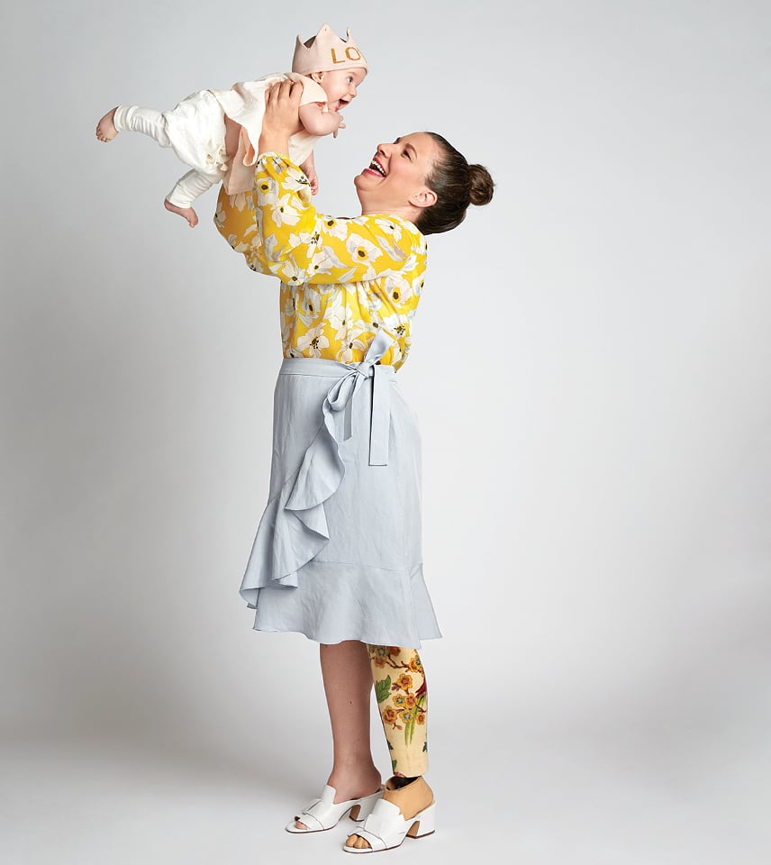 A mom in a floral blouse and skirt holds up her baby while smiling