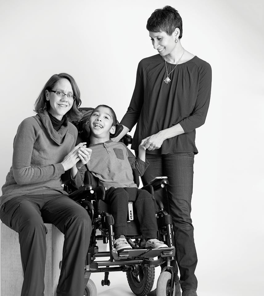 Two women stand close to little boy in a wheelchair that they both care for