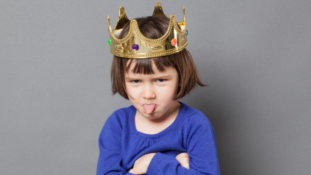cheeky preschool child with golden crown on head folding arms and sticking out tongue