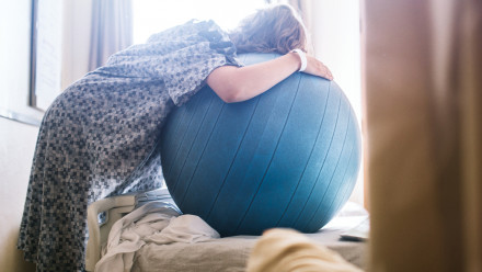 A woman in lobourwith contractions prepares to give birth in a clean white hospital setting. She leans on a birthing or fitness ball to ease the intensity of her contractions.