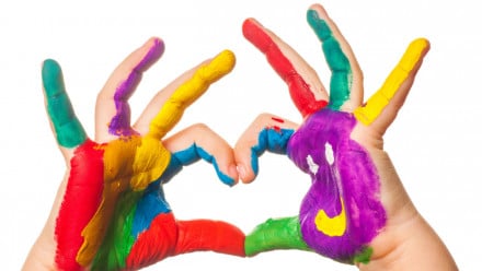 Kid making a heart shape with his fingers, with colourful painted hands.