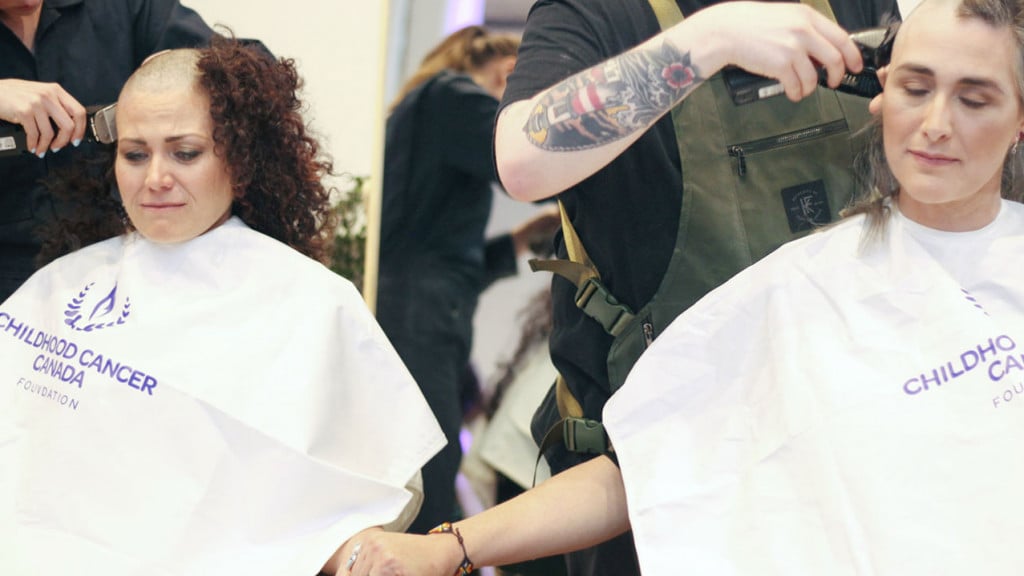 Two women sitting and holding hands as they get their hair shaved 