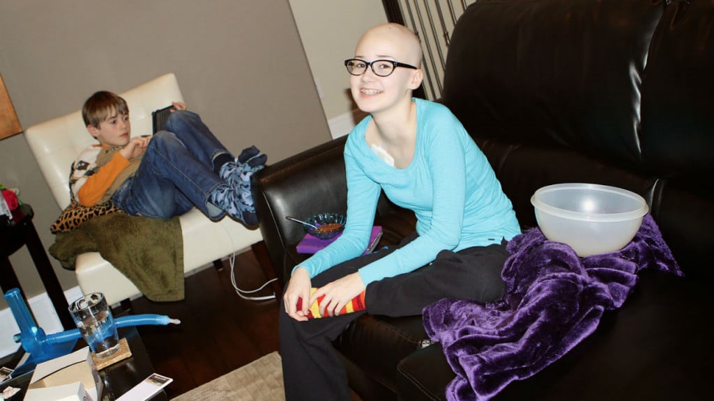 A young girl, suffering from cancer sitting on a couch in her home with no hair 